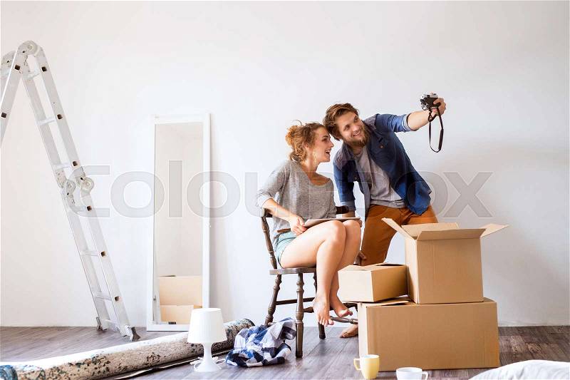 Young married couple moving in a new house, woman with tablet sitting on chair, man taking selfie of them, stock photo