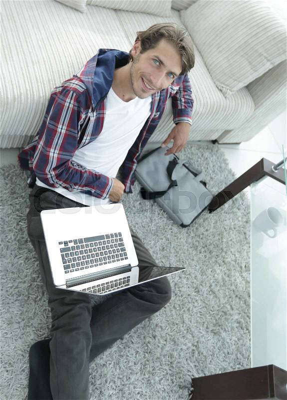 Business guy with laptop sitting near sofa on carpet in living room, stock photo