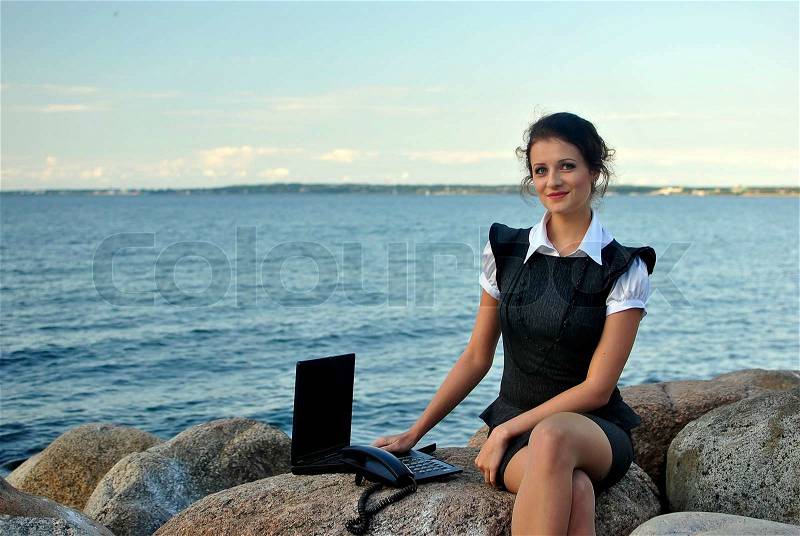Office by the sea: the girl on the rocks with a laptop and a desktop phone, stock photo