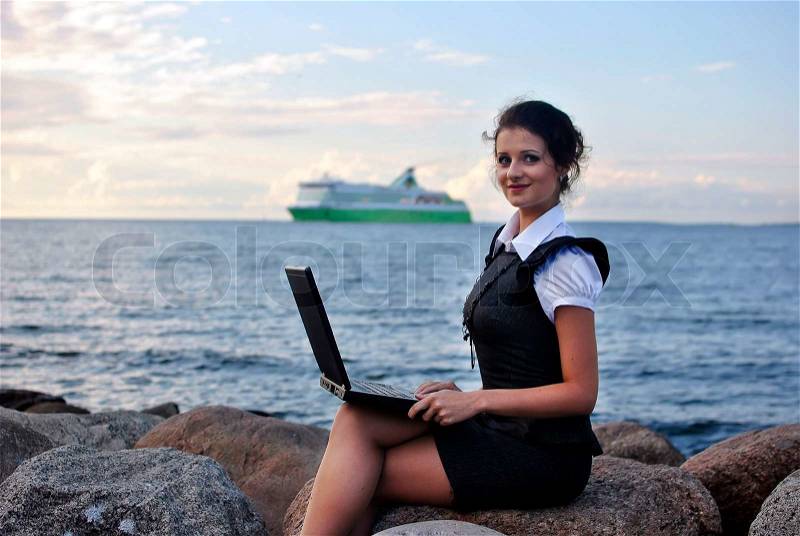 Office by the sea: the girl on the rocks with a laptop ship on the backdrop, stock photo