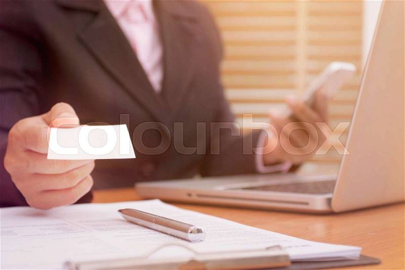 Business woman holding white blank business card and using mobile smart phone at office, stock photo