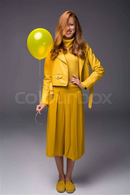 Attractive redhead fashionable girl in yellow clothes with balloon, on grey, stock photo