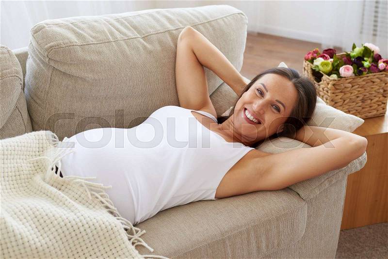 Side view of happy pregnant woman lying down on sofa with hands behind her head, stock photo