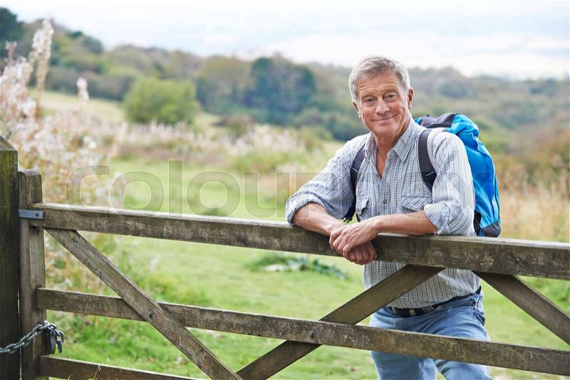 Portrait Of Senior Man On Hike In Countryside Resting By Gate, stock photo