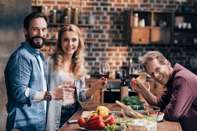 Cheerful middle aged friends drinking wine while cooking dinner and smiling at camera, stock photo
