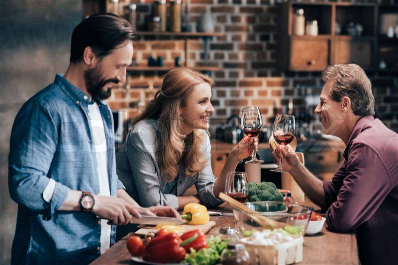 Happy friends drinking wine and smiling each other while man cutting vegetables for salad, stock photo
