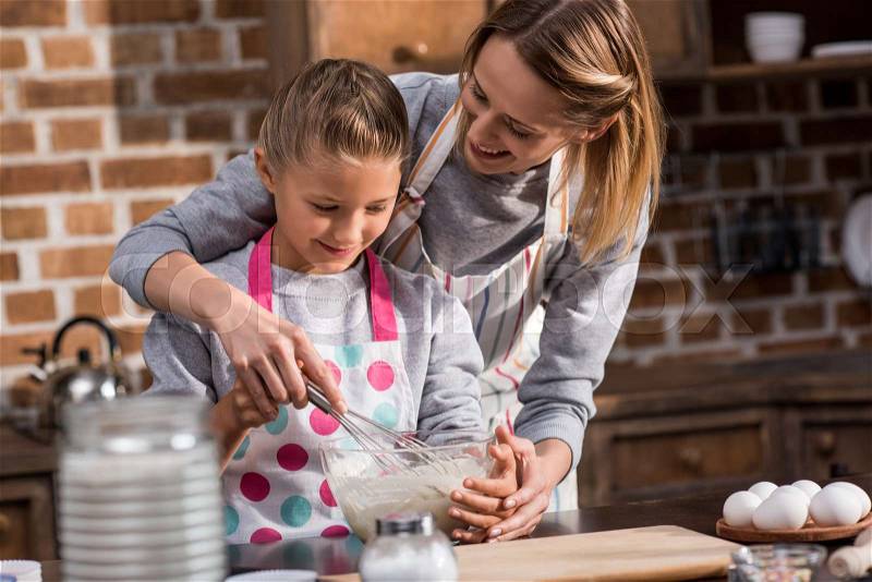 Mother helping little daughter while making cookies together at home, stock photo