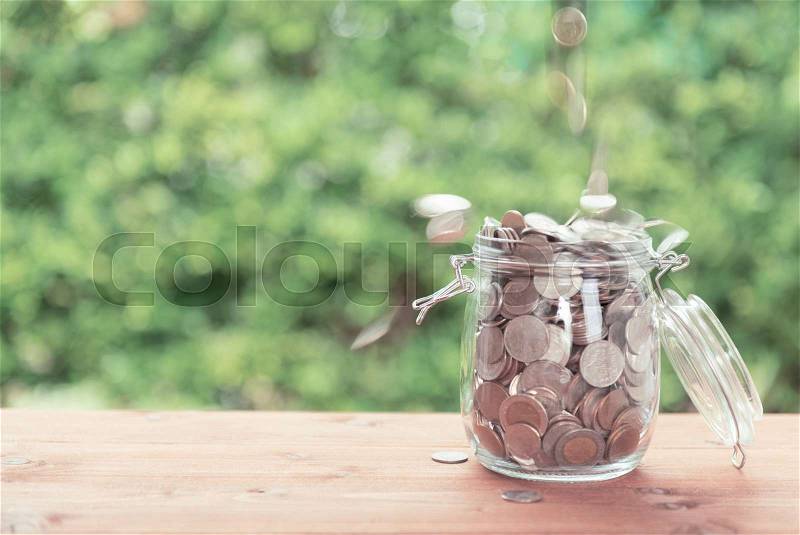 Money coins falling in glass jar on old wooden table with green nature background,saving money concept, stock photo