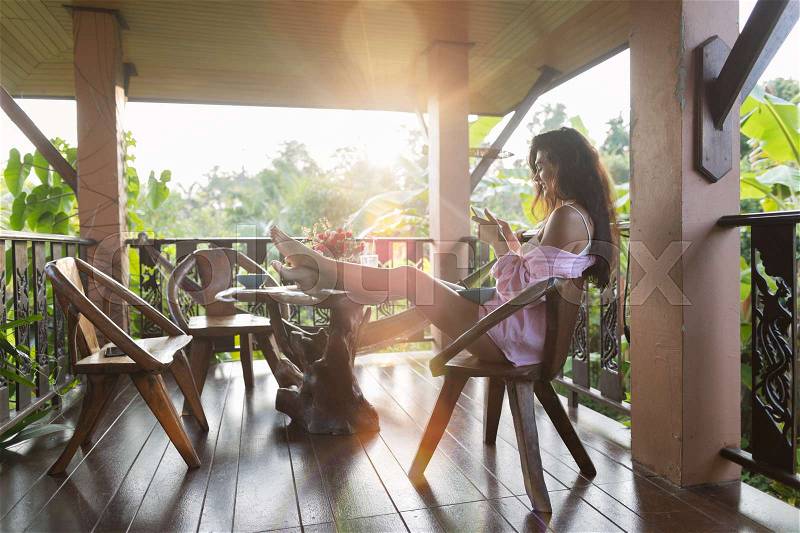 Young Beautiful Woman Using Cell Smart Phone Sit On Terrace With Tropical Forest Landscape And Morning Sun Shine Beautiful Girl Messaging Online Outdoors, stock photo