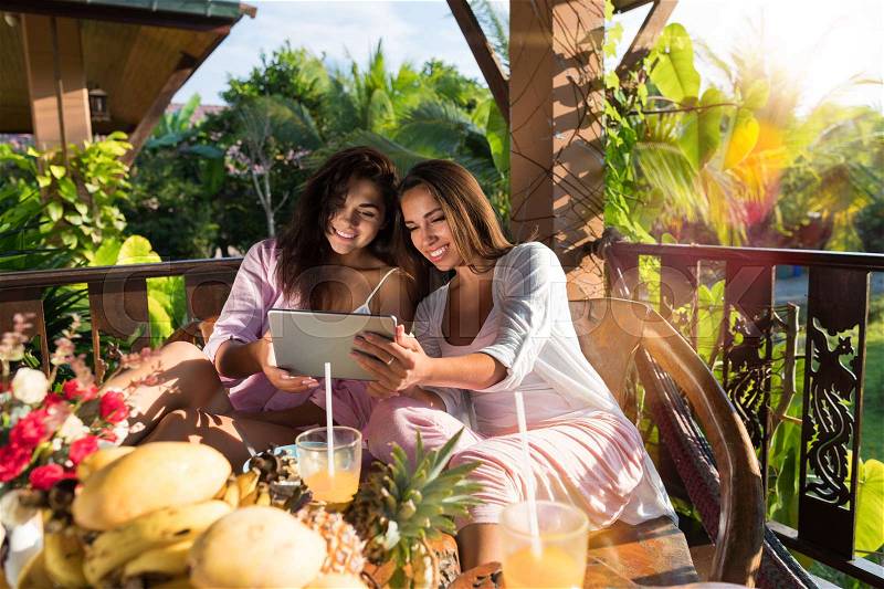 Two Young Women Use Digital Tablet While Breakfast On Terrace In Tropical Garden Beautiful Girls Hold Wireless Mobile Pc Sitting Together At Table Watch Photos In Morning, stock photo