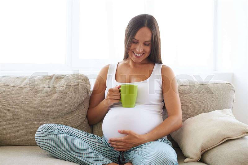 Closeup of pregnant happy woman sitting on sofa and drinking tea, stock photo