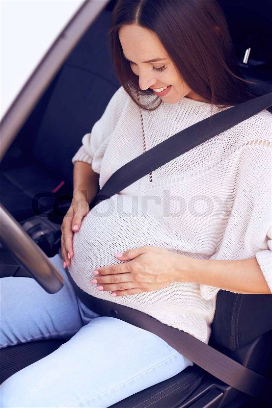 Closeup of excited future mother holding hands on belly in driver seat, stock photo