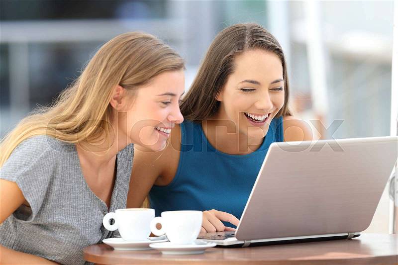 Two happy friends laughing watching on line content in a laptop sitting in a restaurant terrace, stock photo