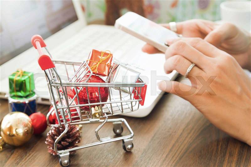 Online payment, Close up at gift boxes in shopping cart and Christmas decoration, woman hands using smartphone and laptop computer in background, stock photo