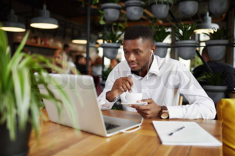 Handsome african freelancer man drinking coffee working at cafe, with laptop. Young male listening music, reading, chatting and watching at gadget. Coffee break of freelancer. Stylish loft interior, stock photo
