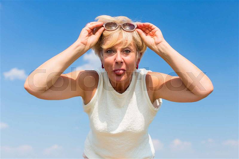 Portrait of a funny senior woman sticking her tongue out while posing outdoors against blue sky in a sunny day of summer, stock photo