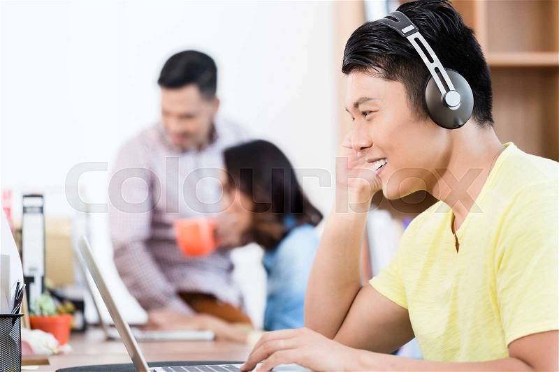 Side view portrait of a happy young Asian employee using headphones while watching a video presentation on laptop in the office, stock photo