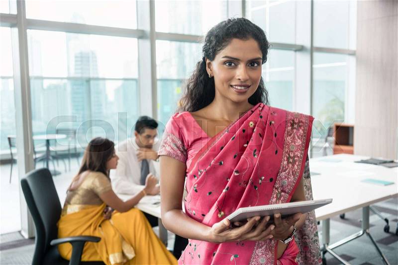 Confident Indian business woman looking at camera while holding a tablet in a modern meeting room, stock photo
