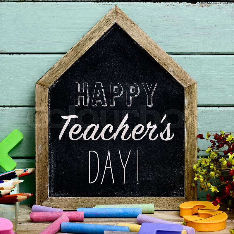 A house-shaped chalkboard with the text happy teachers day written in it on a rustic wooden table full of pieces of chalk of different colors, stock photo