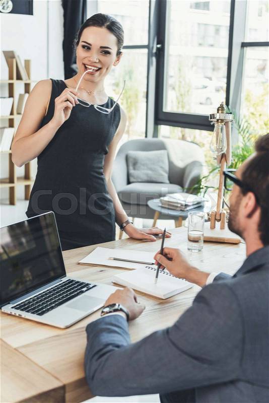 Young businesswoman in black dress standing over desk and smiling flirtatiously at her boss, stock photo