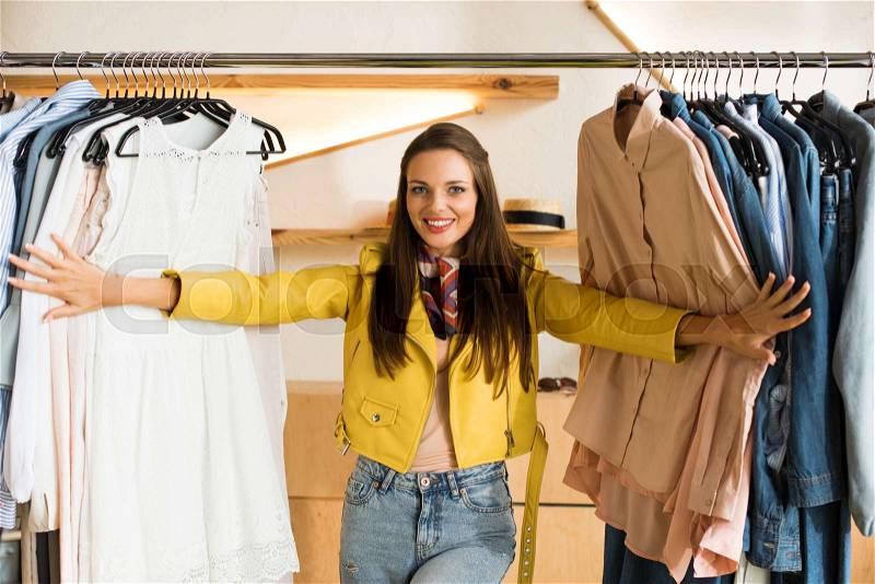 Beautiful young woman choosing clothes and smiling at camera in boutique, stock photo