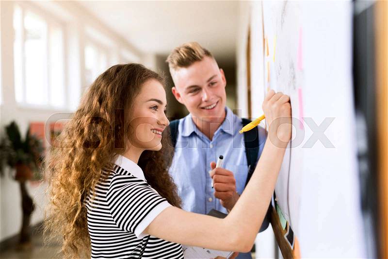 Attractive teenage student couple in high school hall writing something on notice board, stock photo