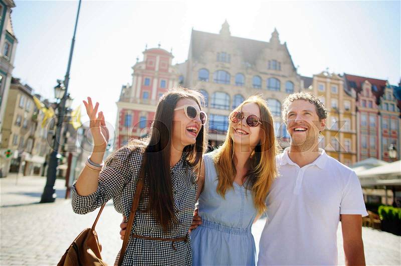 Three young people stand on the square of the ancient city. They are looking at beautiful buildings with pleasure. Friends are hugging each other. Everyone is in a good mood, stock photo