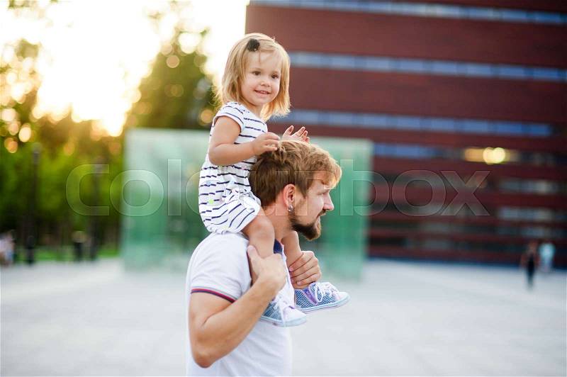 Young man holds his small daughter on his shoulders. The child is smiling. Warm summer day, stock photo