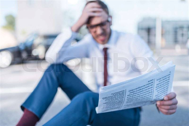 Selective focus of stressed businessman with newspaper sitting on parking with car behind, stock photo