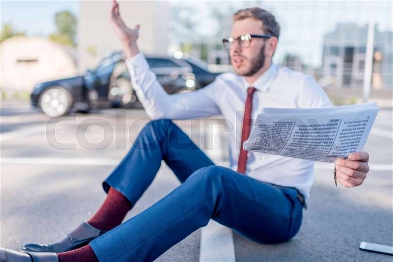 Stressed businessman with newspaper sitting on parking with car behind, stock photo