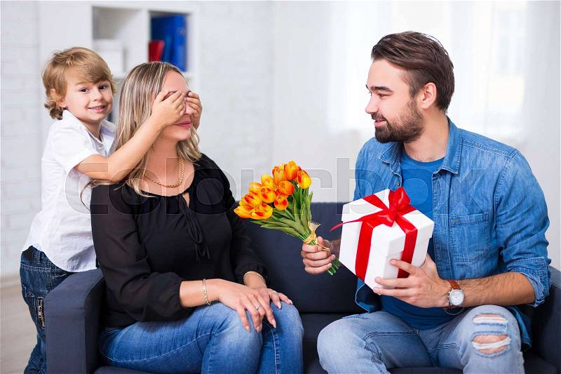 Mother's or female day concept - little son and father giving flowers and gift to wife and mother at home, stock photo