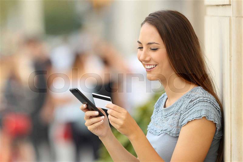 Happy girl paying on line with a credit card and a smart phone leaning on a wall on the street, stock photo
