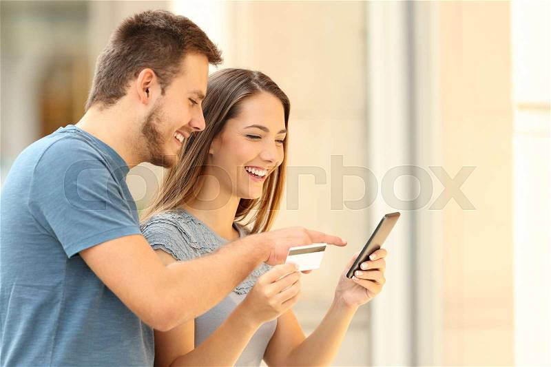 Happy couple buying on line with a smart phone and credit card in front of a storefront on the street, stock photo