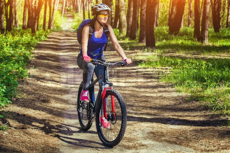 One young woman - an athlete in a helmet riding a mountain bike outside the city, on the road in a pine forest on a summer day, stock photo