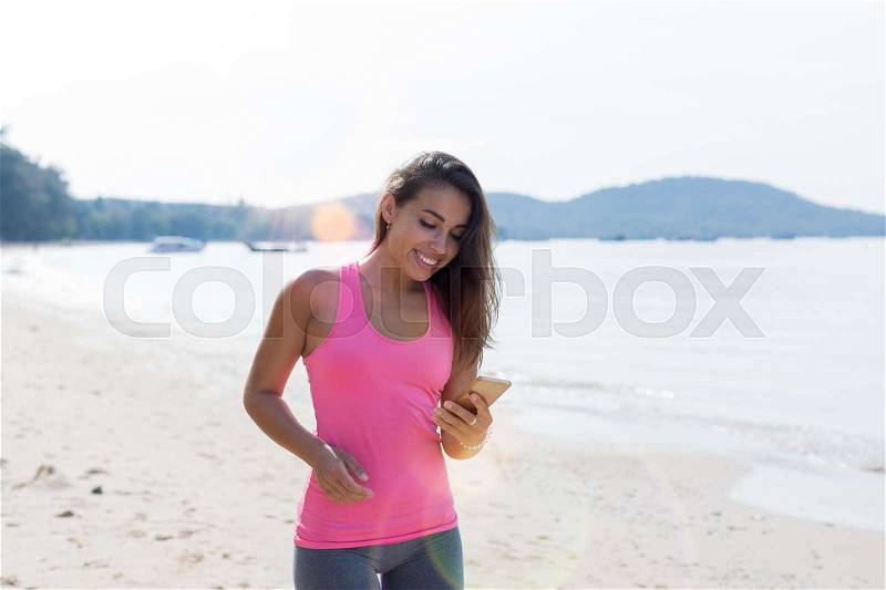 Portrait Of Young Woman Runner On Beach Holding Cell Smart Phone While Jogging On Seaside Female Happy Smiling Girl Sport Runner Healthy Lifestyle Concept, stock photo