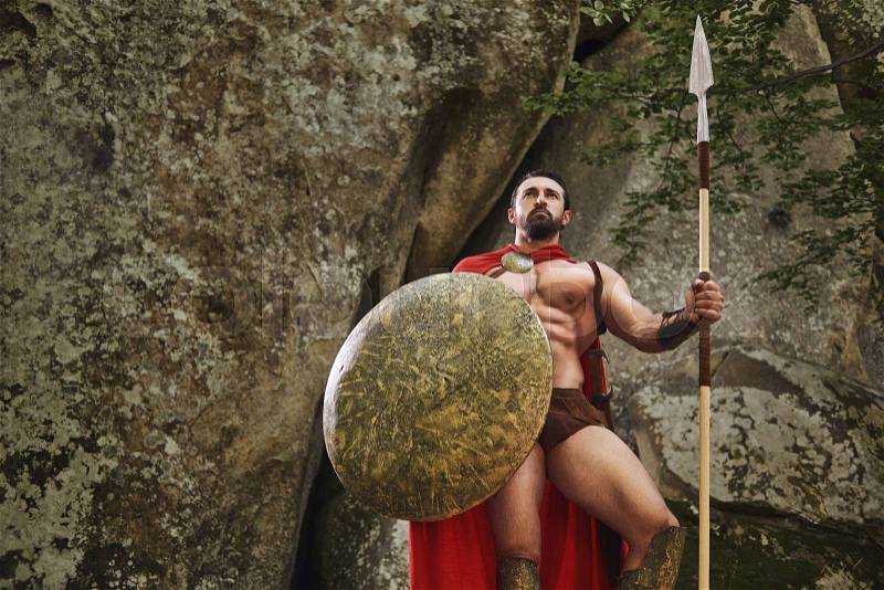 Muscular athlete wearing armor of Roman gladiator and holding shield with spear while posing confidently on background of rocks. Spartan, stock photo