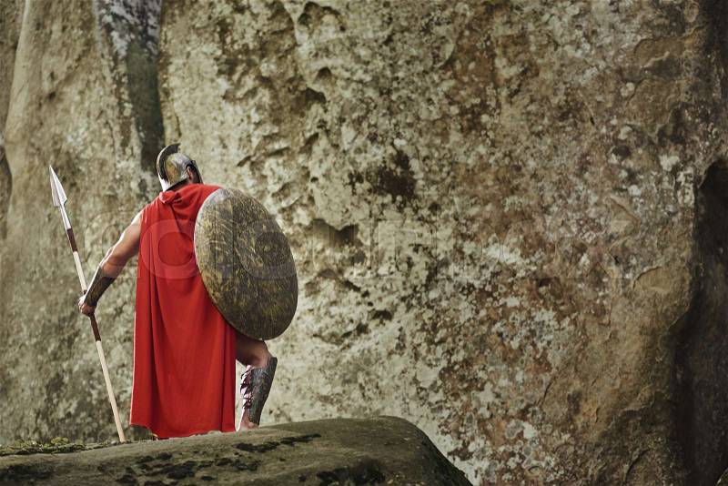 Back view of victorious warrior in long red cloak and gladiator helmet posing with heavy shield and spear standing on rocks. Spartan, stock photo