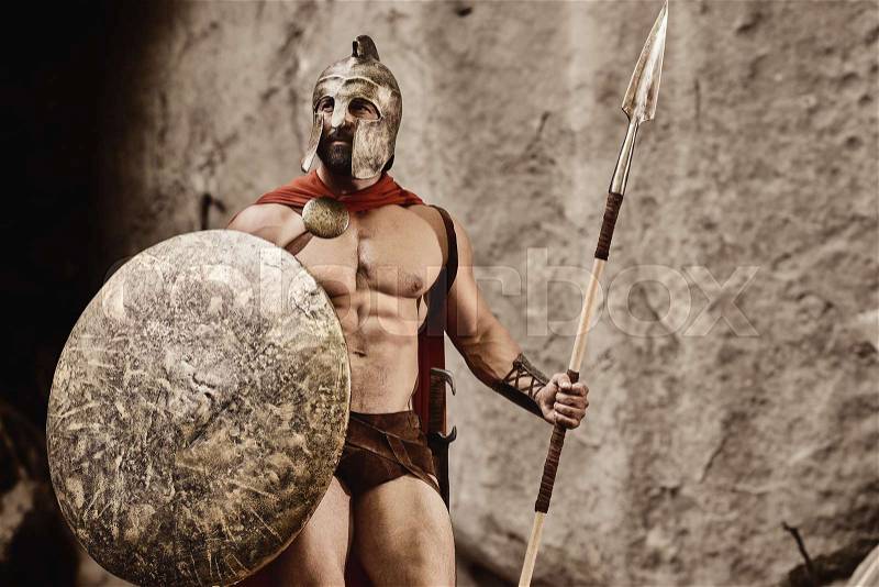 Confident young man with beard and muscular body wearing gladiator armor standing with metal shield and spear on background of rocks, stock photo