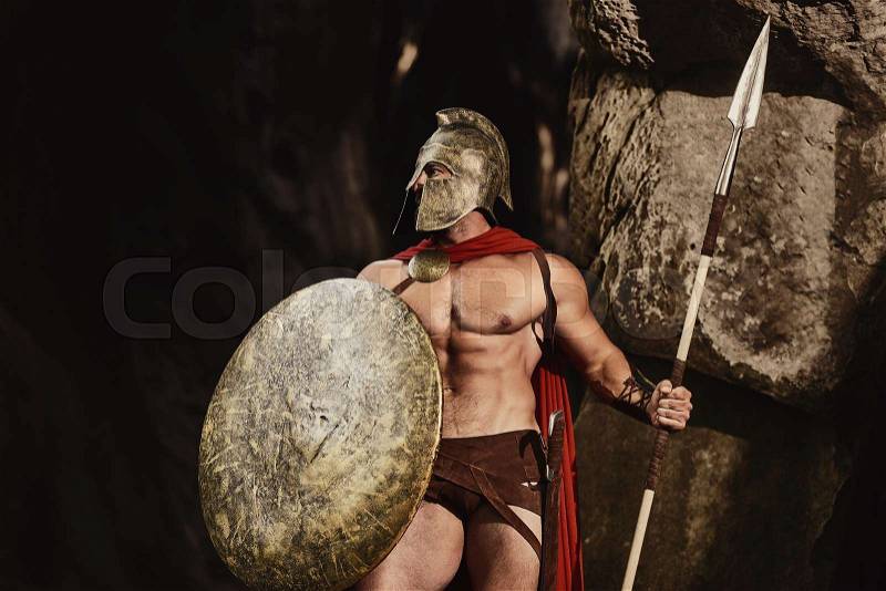 Muscular confident man in helmet and armor of ancient Roman warrior holding spear and shield looking confidently away on nature. Spartan, stock photo