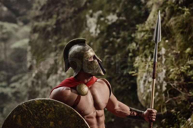 Strong muscular Roman gladiator in helmet and red cloak posing shirtless with shield and spear looking confidently away. Spartan, stock photo
