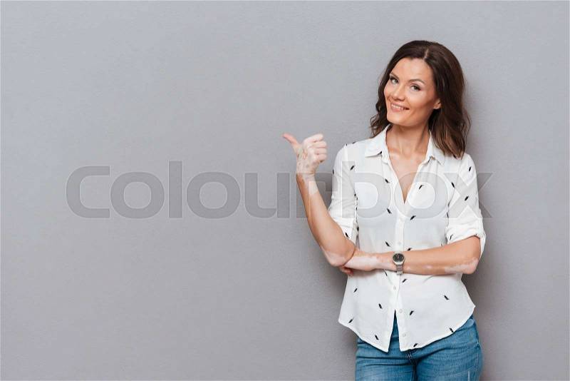 Happy woman pointing away and looking at the camera over gray background, stock photo
