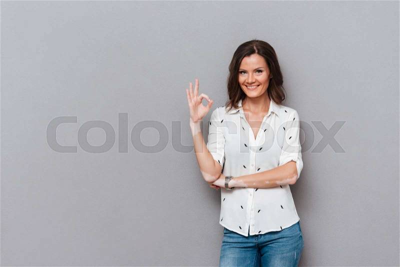 Happy woman showing ok sign and looking at the camera over gray background, stock photo
