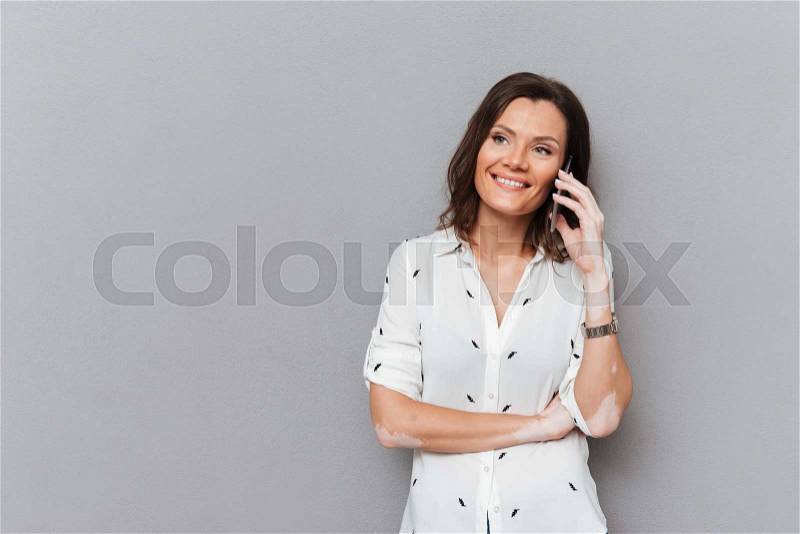 Happy woman talking by the smartphone and looking away over gray background, stock photo