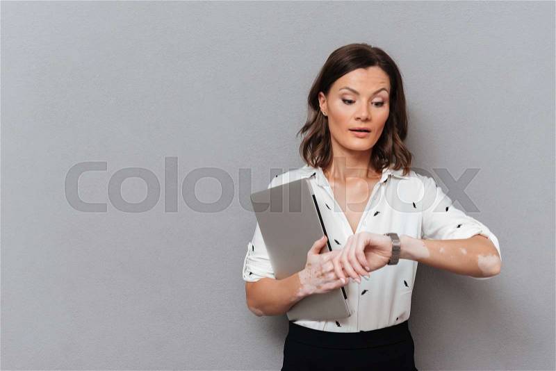 Surprised woman in business clothes with laptop computer in hand looking on wristwatch over gray background, stock photo