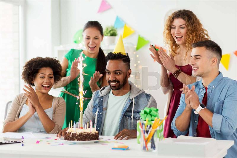 Corporate, celebration and people concept - happy team with firework on birthday cake and non-alcoholic drinks greeting colleague at office party, stock photo