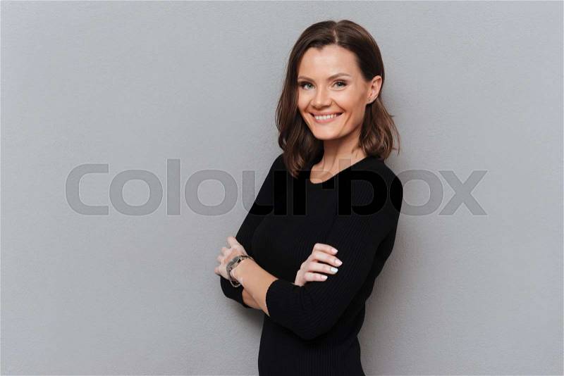 Pleased woman in black dress posing with crossed arms and looking at the camera over gray background, stock photo