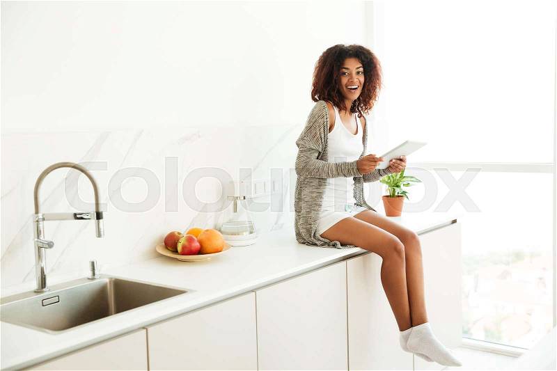 Excited happy afro american woman using pc tablet while sitting on a kitchen table at home and looking at camera, stock photo