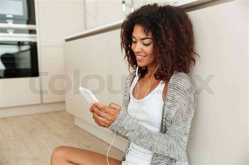Side view of a smiling african woman sitting on the floor in kitchen at home and using smartphone, stock photo