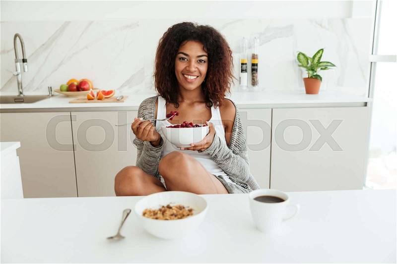 Beauty happy african woman eating by the table in kitchen and looking at the camera, stock photo