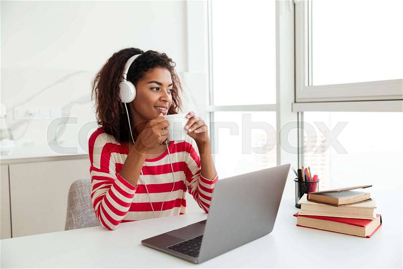 Smiling african woman sitting by the table with laptop computer and drinking coffee, stock photo